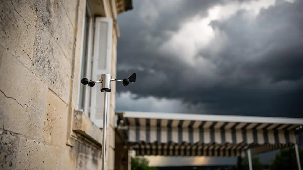 Petit capteur de vent monte sur la facade d'une maison francaise, sentinelle discrete face a un ciel d'orage menacant au-dessus d'un store banne replie